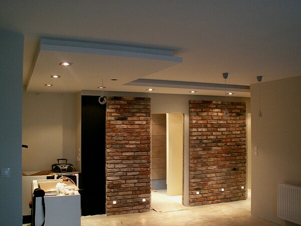 Wykończone wnętrze z dekoracyjną ścianą z cegły klinkierowej z punktowym oświetleniem u dołu, widoczne fragmenty sufitu podwieszanego z halogenami i wiszące lampy.